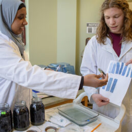 Two scientists in lab coats are collaborating in a laboratory setting. One is a woman wearing a hijab and the other is a man with long hair. They are examining test results. The workspace includes jars filled with a dark liquid and other lab equipment.