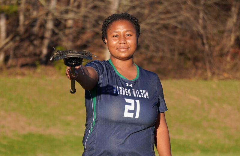 Madison Sings, a senior at Warren Wilson College, stands in her lacrosse jersey and points her lacrosse stick to the camera.