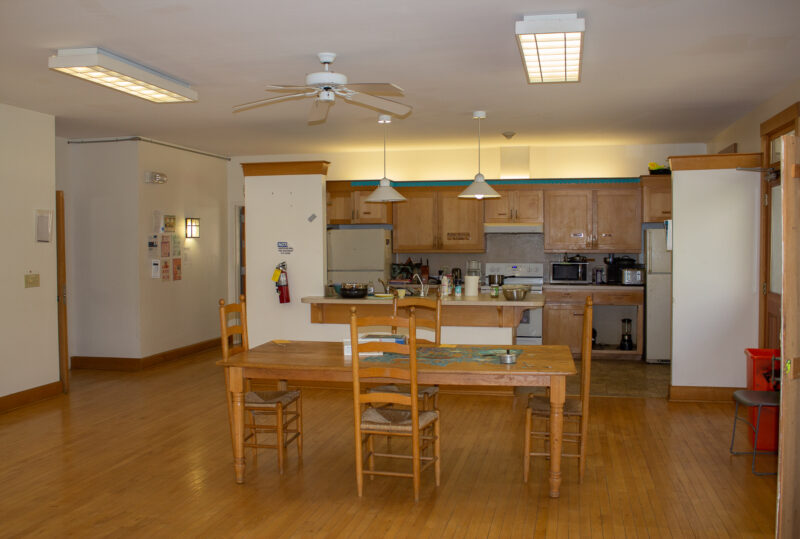 The Wellness dorm kitchen. A dining room table is in the middle room chair. On the far wall there are two refrigerators, a stove, a microwave, a coffee pot, and blender.