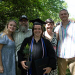 A graduate stands with 4 other people holding their pine tree and degree smiling.