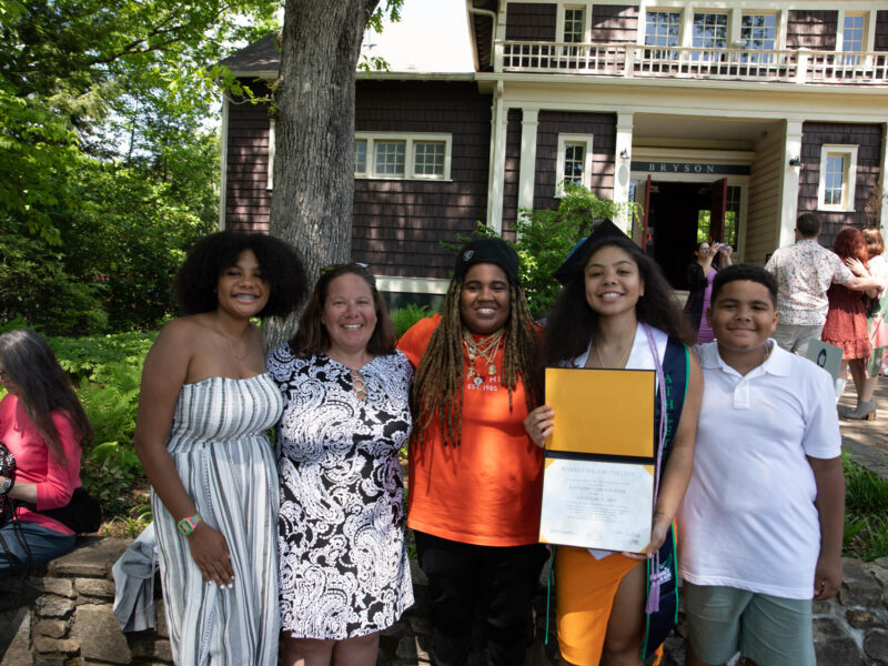 A graduate is joined by four people in a photo. They are all smiling. The Graduate is showing their degree.