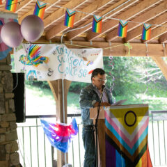 Director of the Queer Resource Center, Jo Go, speaks to those gathered at the lavender graduation. Various Pride flags are hung up in the background.