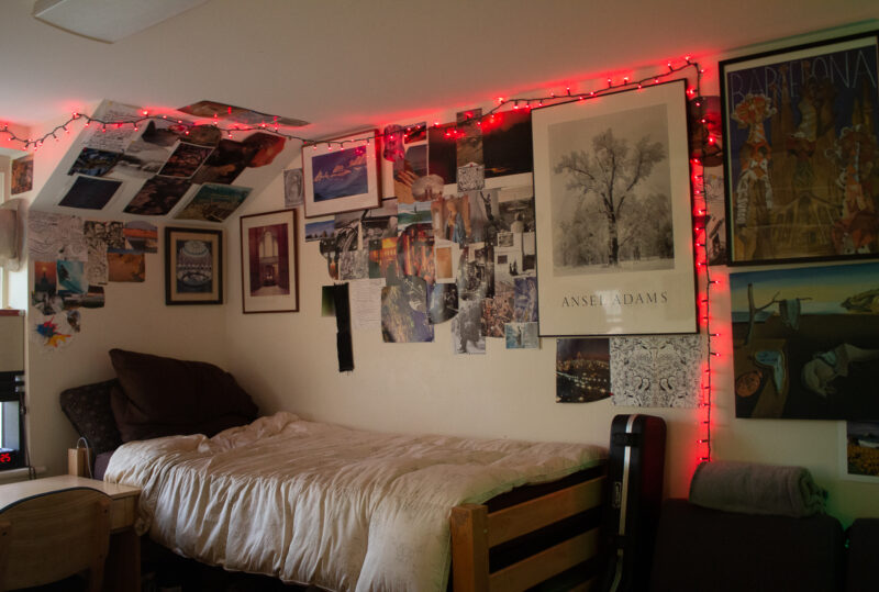 A dorm room with a desk and bed. The walls around the bed are covered in pictures and lights.