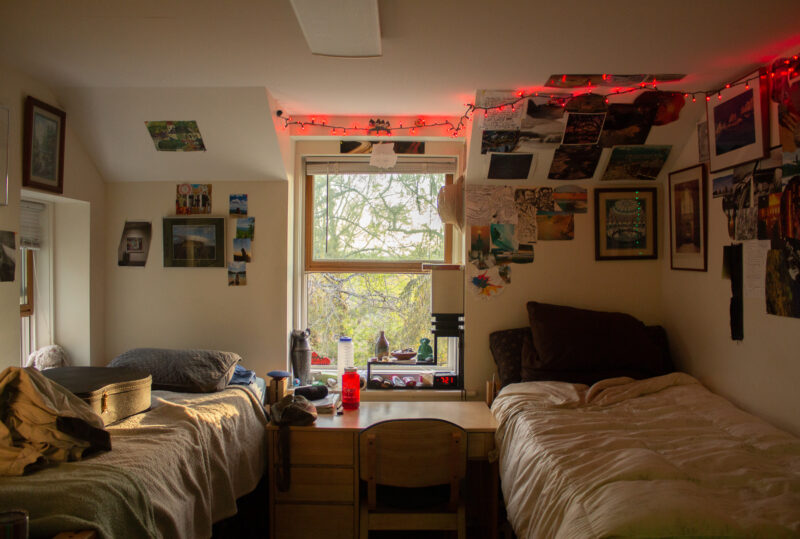 A dorm room in sage. There are two beds on either side of the room with a desk in the middle and a window above the desk. The walls are covered in posters, pictures, and lights.