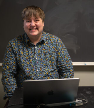 Communications Professor Beck Banks sits at a desk with a macbook smiling
