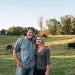 Alumni Jamie and Amy Ager stand smiling in front of cows at their farm Hickory Nut Gap