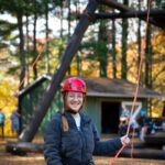 Student Ahna Webster stands at the base of the Alpine Tower challenge course holding ropes and smiling.