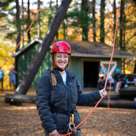 Student Ahna Webster stands at the base of the Alpine Tower challenge course holding ropes and smiling.