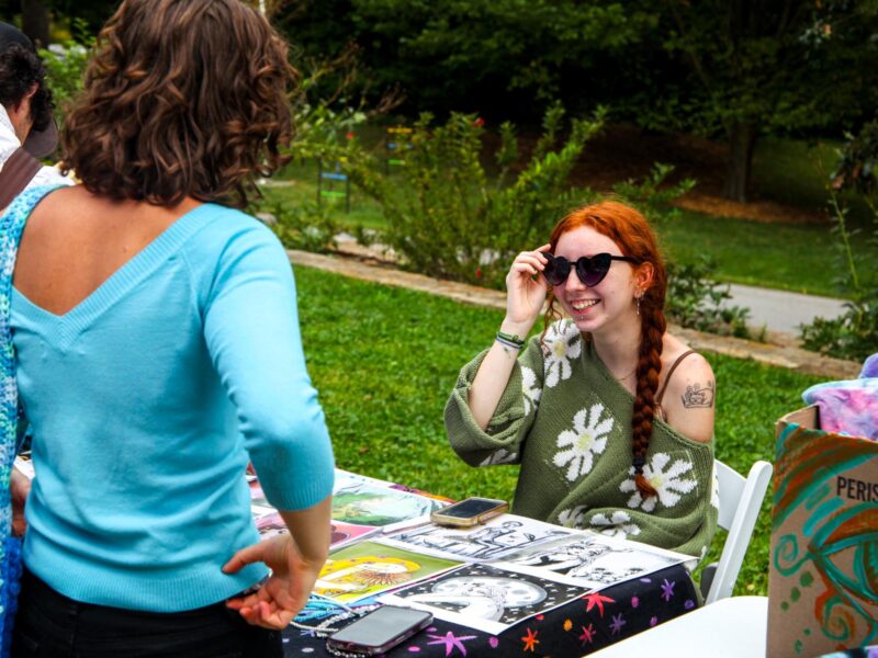 A student smiles from behind a table of their art work they are selling at a craft fair.