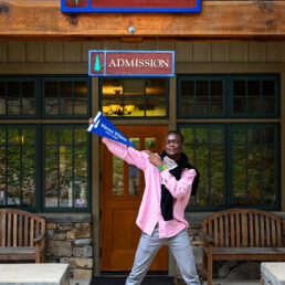 Student Munashe stands smiling and holding a warren wilson pennant in front of the admissions office.