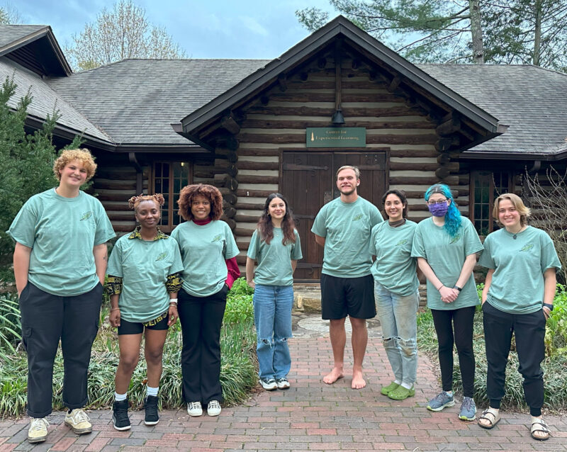 8 students wearing blue ecoteam shirts stand smiling in front of the log cabin on campus.