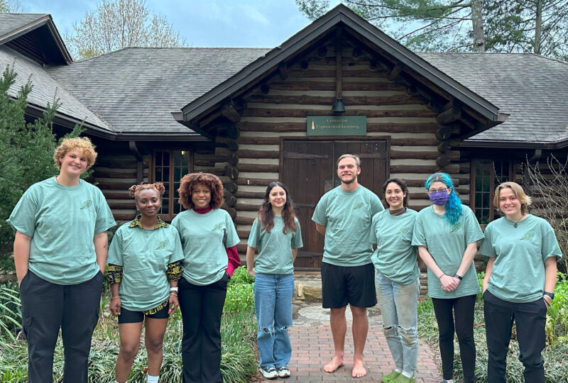 8 students wearing blue ecoteam shirts stand smiling in front of the log cabin on campus.