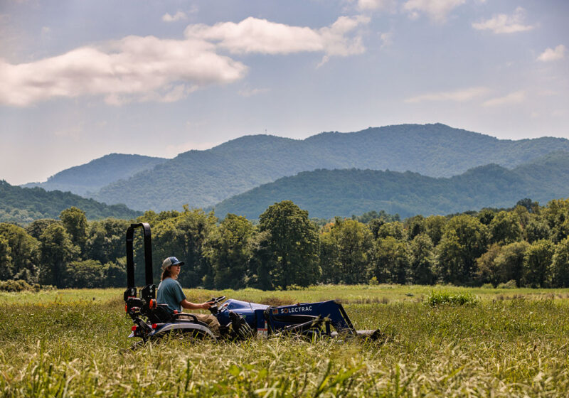 A student drives an Electric Tractor through a pasture with a beautiful mountain range in the background.