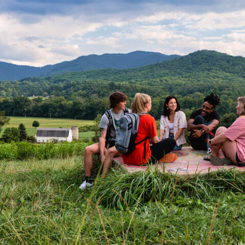 A group of students sit on a concrete pad smiling and laughing. Behind them is a beautiful mountain vista and the white barn.