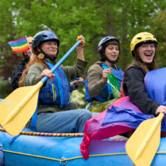 A group of adventure programs students sit on a whitewater raft that has been turned into a parade float for the Big Gay Earth Day Parade. They are each holding paddles and pride flags and smiling.