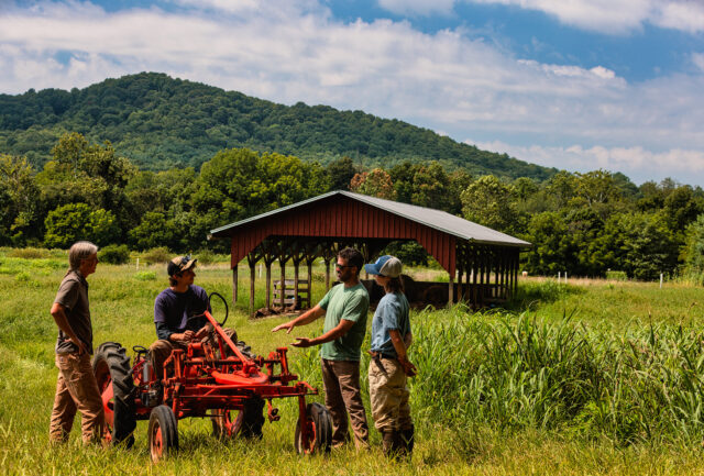 A student and two staff stand around a tractor with another student on it. The tractor is in a field with a red open barn and mountains in the background.