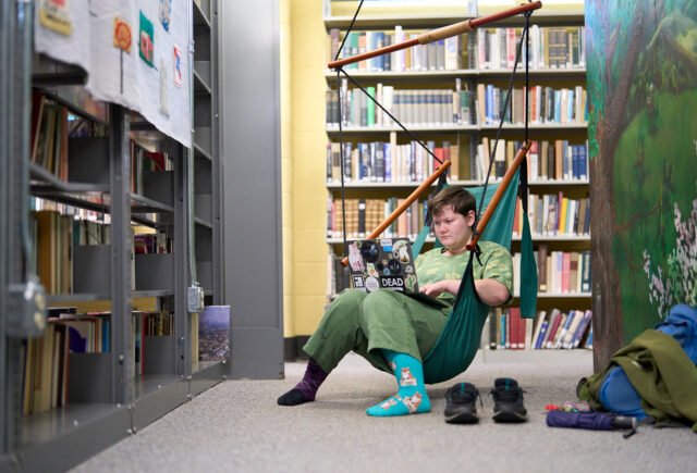 A student sits in a hammock chair in the library studying on their laptop.