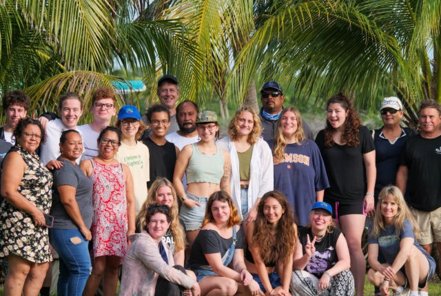 A group of students on a study away trip in Belize pose together for a picture with the back drop of thick palm trees.