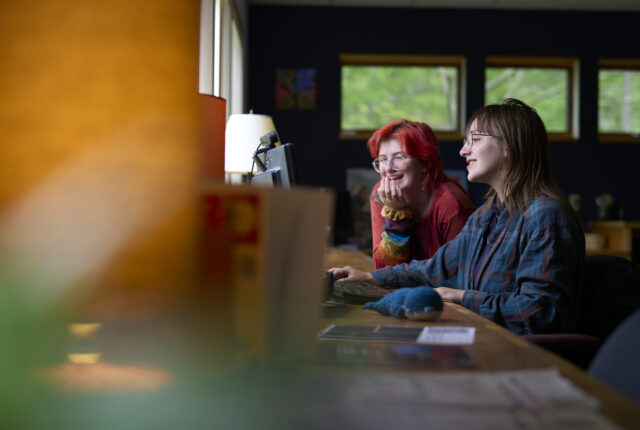 Two students sit at a computer in the writing studio. The student with red hair is laughing and the student with the brown hair looks to be talking