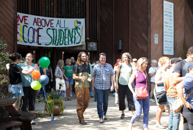 A group of students and their families walk out of the chapel. Above the doors is a large banner that reads `Love above all` `Welcome Students`.