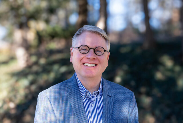 David Gliem stands in front of foliage and smiles for a headshot.